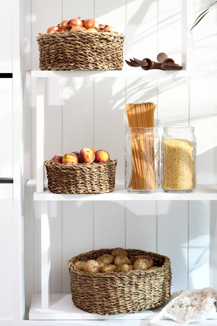 services-02 A neatly organized pantry shelf with wicker baskets holding apples, peaches, and potatoes, alongside pasta and grains.
